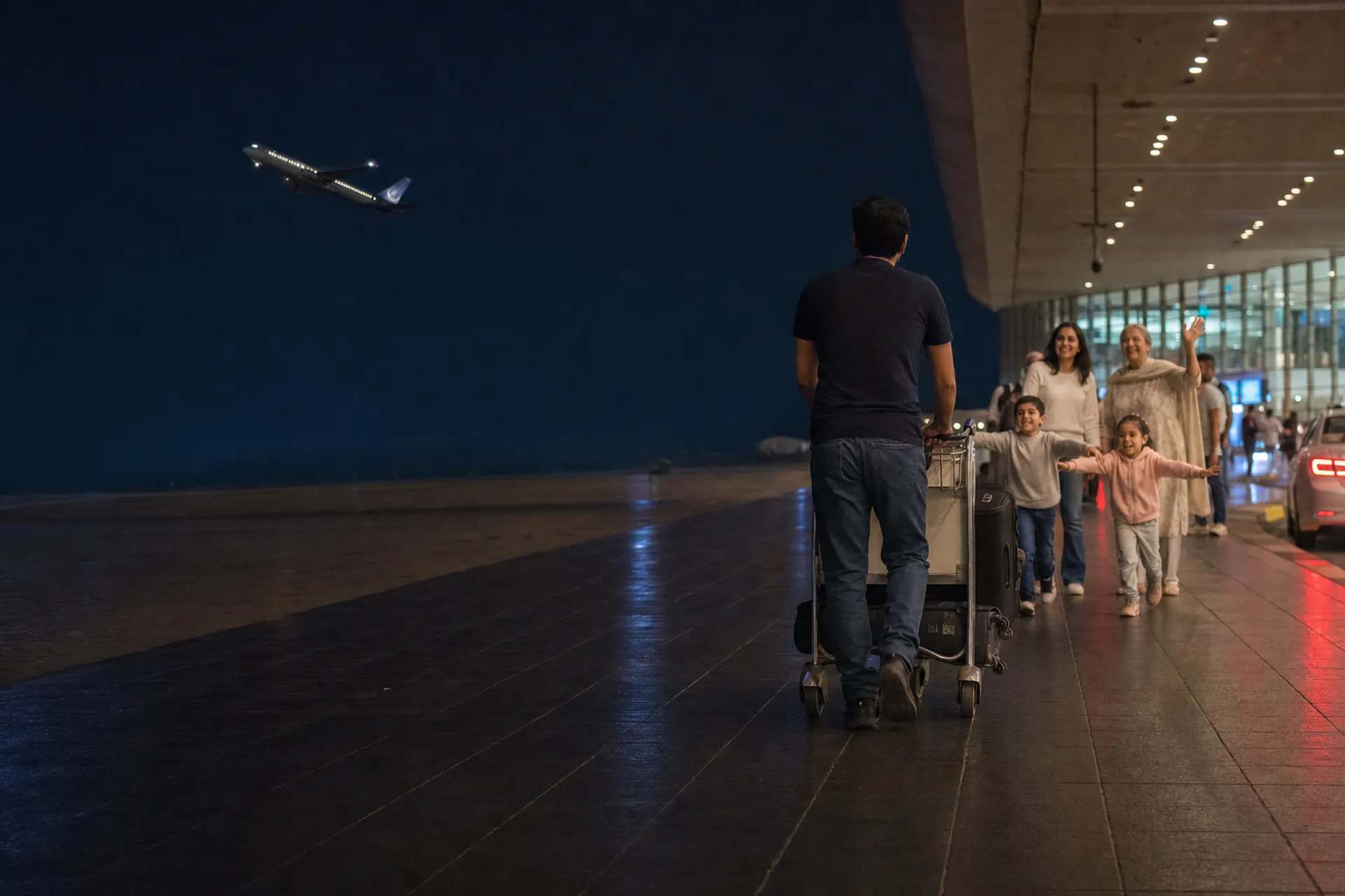 The same airport forecourt after dark: a traveller seen from behind pushing a luggage trolley, his family greeting him on the right with two children running towards him under warm terminal lighting, and a lit airliner on a deep navy night sky to the left.